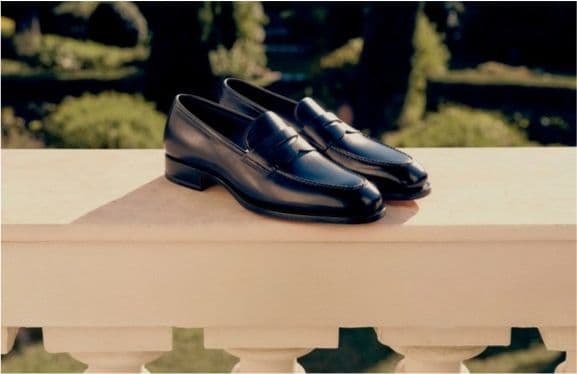 Loafers - Santoni black leather loafers on a stone ledge with formal gardens behind.